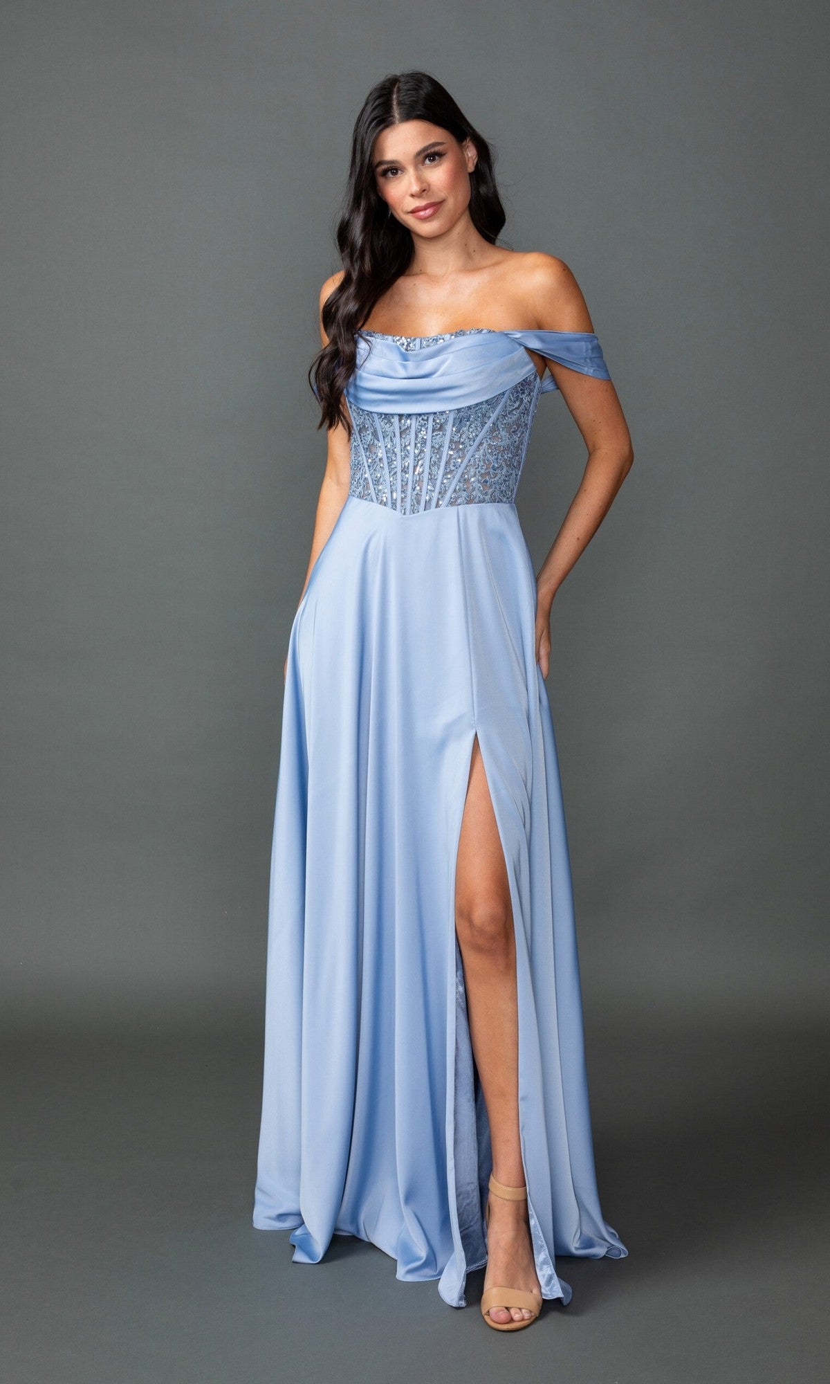 A woman stands against a gray background wearing the Off-Shoulder Corset Prom Gown: Dancing Queen 4546A, featuring an embellished corset bodice, thigh-high slit, and nude heeled sandals.